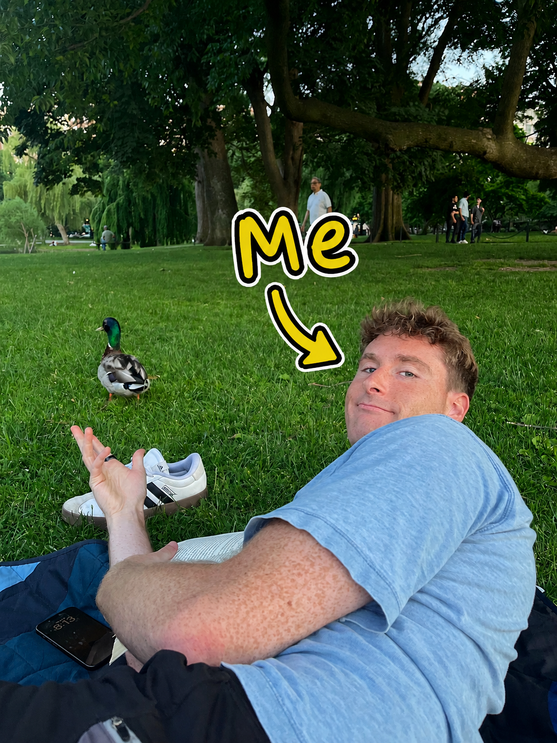 Dan lying in the grass near a duck in a park, with a sticker pointing to him.